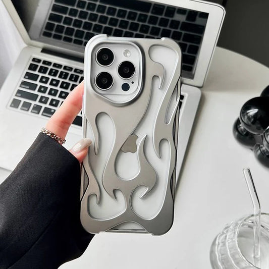 A silver protective case for an iPhone, featuring a flame design, being held by a person with one hand, placed on a desk next to a laptop and a glass.